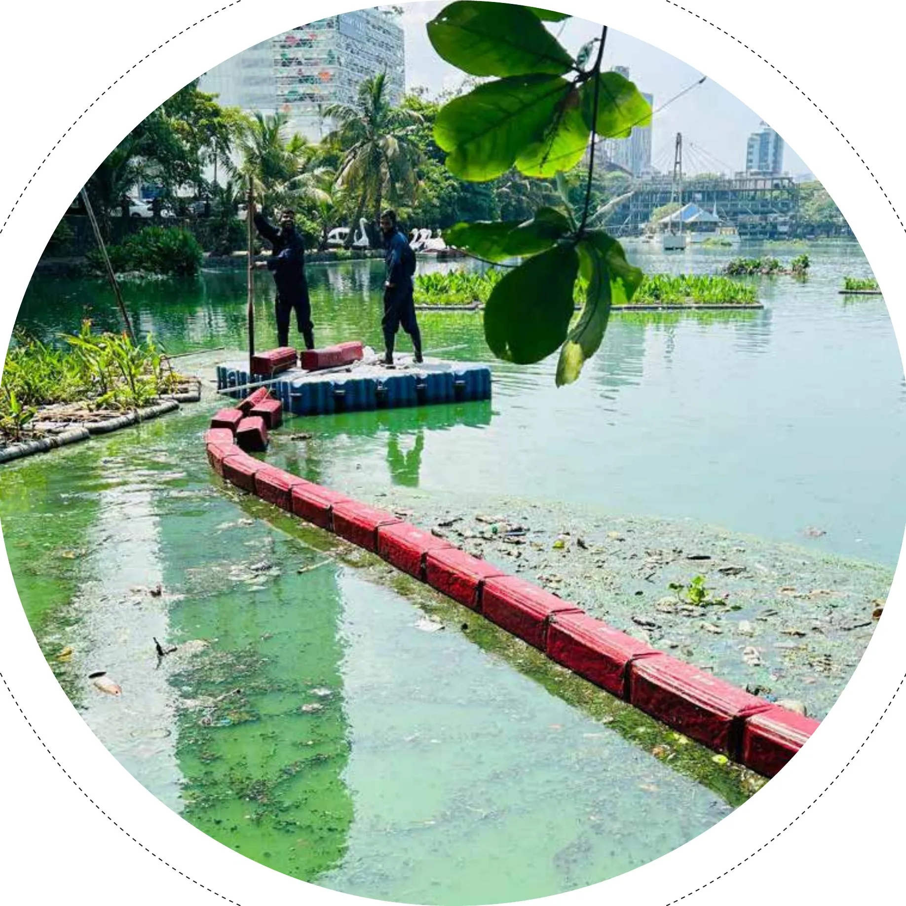 A floating OCEAN STRAINER™ barrier of red and blue pontoon blocks spanning a green, waste-strewn urban waterway, with two workers standing on a floating platform.
