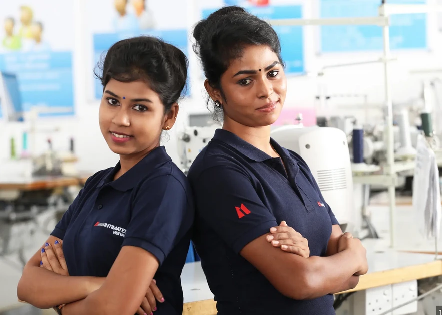 Two MAS women employees in navy polos standing back-to-back with arms crossed on the factory floor