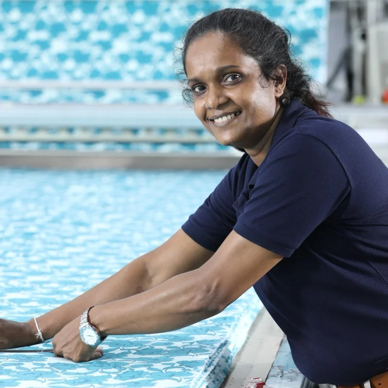 A smiling MAS woman employee in a navy polo leaning by a swimming poolside