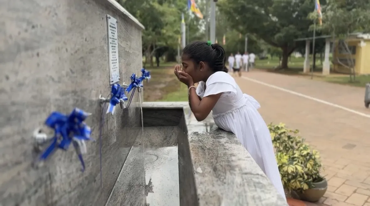 A young girl cupping water from blue ribbon-tied taps at an outdoor communal water station, with a school sports ground behind
